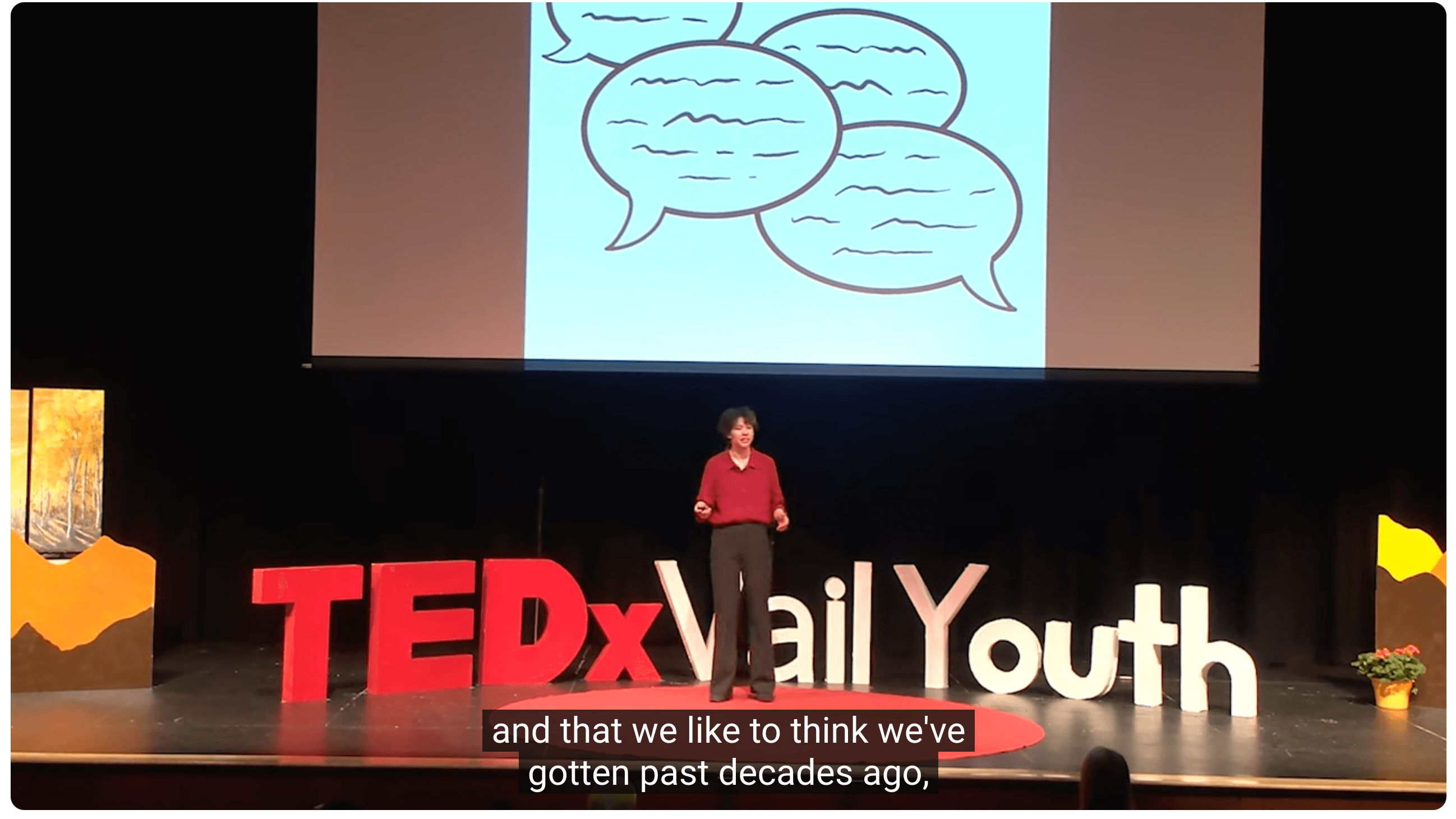 Student in black pants and red shirt stands on stage with conversation bubbles on the screen